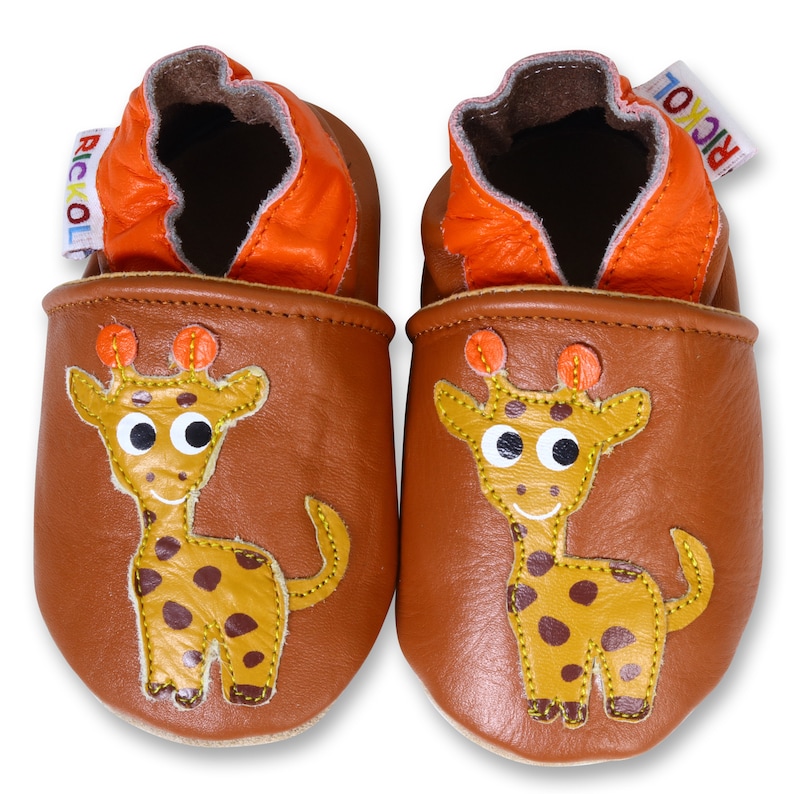 Puede incluir: Un par de zapatos de beb&eacute; de cuero marr&oacute;n con un dise&ntilde;o de jirafa. Los zapatos tienen un forro naranja y una etiqueta Rickol.