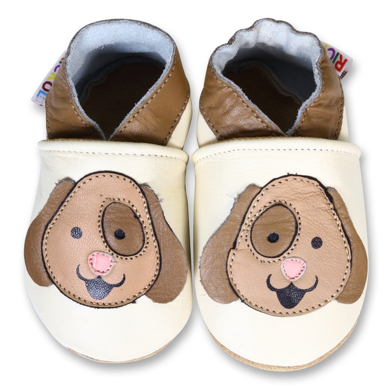 Puede incluir: Un par de zapatos de beb&eacute; de cuero color crema con un dise&ntilde;o de cara de perro marr&oacute;n. Los zapatos tienen un ribete de cuero marr&oacute;n y una suela suave y flexible.