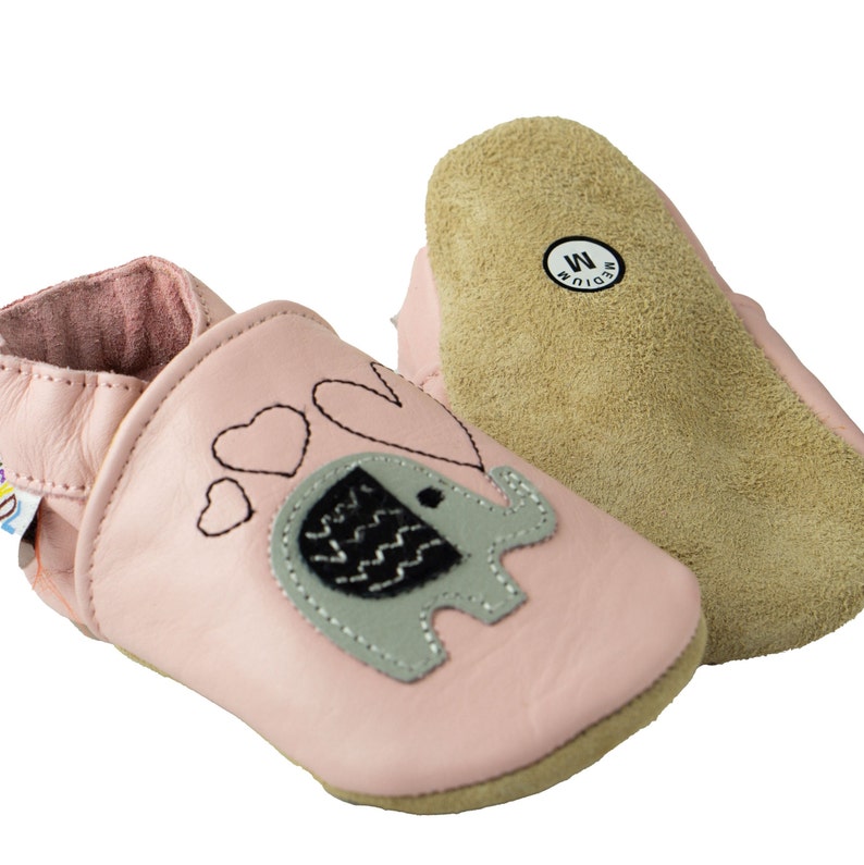 Puede incluir: Zapatos de beb&eacute; de cuero rosa con un dise&ntilde;o de elefante y corazones. Los zapatos tienen una suela de gamuza y dicen "Medium" en la parte inferior.