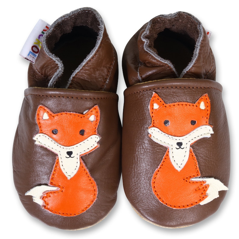 Puede incluir: Un par de zapatos de beb&eacute; de cuero marr&oacute;n con un dise&ntilde;o de zorro. El zorro es naranja con la cara y la cola blancas.