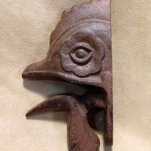 Puede incluir: Un tope de puerta de hierro fundido de color marrón con forma de gallo. La cabeza del gallo está levantada y su pico está abierto. El ojo del gallo es visible y se pueden ver los detalles de las plumas.