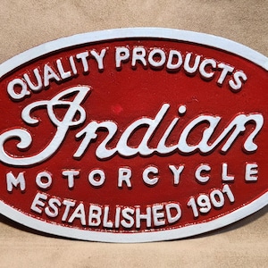 Peut inclure: Une plaque ovale en métal rouge et blanc avec le texte en relief blanc "QUALITY PRODUCTS", "Indian", "MOTORCYCLE" et "ESTABLISHED 1901". La plaque présente une bordure blanche et deux petits trous sur les côtés gauche et droit.