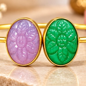 Könnte beinhalten: Ein goldfarbenes Armband in offener Form mit zwei ovalen Cabochons. Ein Cabochon ist hellviolett und der andere leuchtend grün, beide mit einem Blumenmuster graviert. Das Metallband ist glatt und poliert und verbindet die beiden Fassungen.