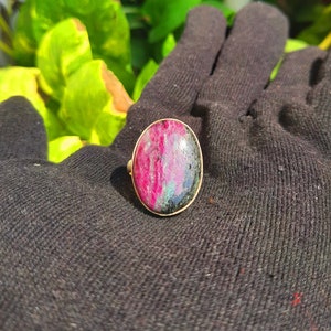 以下が含まれることがあります： 赤、黒、緑の色合いが混ざった、楕円形の宝石をあしらったゴールドトーンのリング。リングはシンプルなポリッシュゴールドバンドで、石の自然な美しさを際立たせています。ジュエリーです。