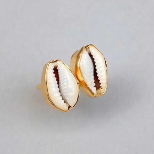 Puede incluir: Un par de pendientes de botón con conchas de cauri blancas naturales engastadas en metal dorado pulido. Cada concha muestra su característica abertura estriada y una superficie curva y lisa, asegurada en un marco de metal de color dorado.