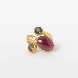 May include: A gold ring featuring three gemstones. The largest stone is oval-shaped and pink, while the two smaller stones are round and brown.