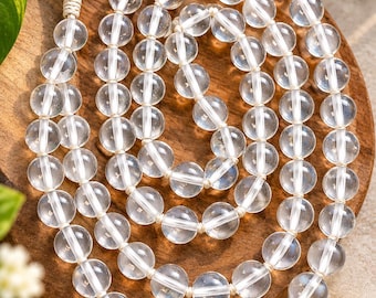 Puede incluir: Un largo collar de cuentas de cuarzo esféricas y transparentes dispuestas en un patrón circular, con una borla de tela de color crema en la parte superior. Las cuentas están ensartadas en un cordón claro, formando un bucle continuo de piedras pulidas.