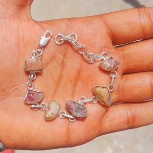 Peut inclure: Un bracelet en argent orné de pierres précieuses brutes de forme irrégulière, dans des tons de rose, pêche et jaune. Les pierres sont serties dans des chatons en argent et reliées par une chaîne et un fermoir. Le bracelet est présenté sur une main.