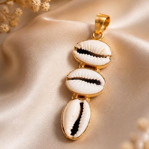 Pode incluir: Um pingente de tom dourado com três conchas de búzios brancas dispostas verticalmente em uma fileira. Cada concha está inserida em uma moldura de metal dourado, com uma argola dourada no topo para prender a uma corrente. As conchas possuem estrias naturais e aberturas escuras.