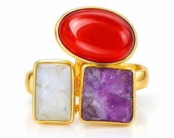 May include: A gold-toned ring featuring three distinct stones: a large, smooth, oval-shaped red stone positioned above a rectangular, textured white stone and a square, rough-cut purple stone. The metal band is polished and gold in color.