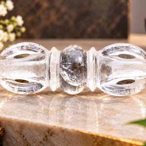 Puede incluir: Un vajra de cristal de cuarzo transparente con una cuenta central esférica flanqueada por dos extremos simétricos de múltiples puntas. El objeto está tallado en una sola pieza de piedra natural con inclusiones internas visibles.
