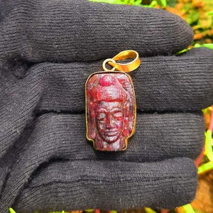 May include: A rectangular pendant featuring a deep red carved gemstone depicting a human face, set in a gold-toned metal frame with a matching gold-toned bail at the top. The pendant is held against a dark, textured fabric background.
