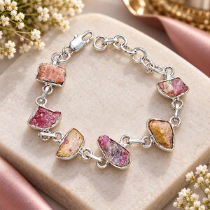 May include: A silver chain bracelet featuring six irregular, raw gemstone segments in shades of pink, magenta, and golden yellow. The bracelet is constructed with polished silver links and includes a secure lobster claw clasp closure mechanism.