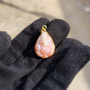 May include: A teardrop-shaped pendant with a gold-coloured frame. The pendant features a carved, light pink and white figure. The pendant is held by a black glove. The pendant is a jewelry item.