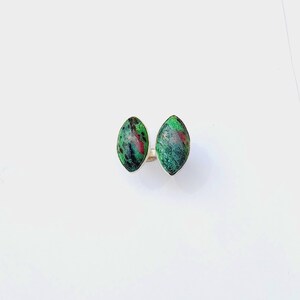 Puede incluir: Un anillo de plata con dos piedras preciosas ovaladas. Las piedras preciosas muestran una mezcla de colores verdes, rojos y negros. El anillo tiene un diseño sencillo, con las piedras colocadas una al lado de la otra sobre una banda de plata. Las piedras preciosas tienen un aspecto natural y texturizado.