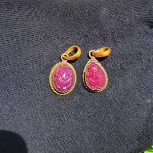 May include: Two gold pendants with a carved pink gemstone. The pendants are shaped like teardrops and have a detailed design.