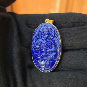 Puede incluir: Colgante ovalado con una figura azul tallada en un marco dorado. El colgante presenta una talla detallada de una figura sentada con un halo. El marco dorado tiene un diseño simple y elegante. El colgante está sobre un fondo negro.