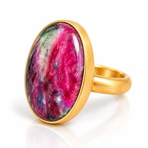 Puede incluir: Un anillo de oro con una gran piedra preciosa en forma de cabujón ovalado con una superficie pulida. La piedra muestra una mezcla vibrante de inclusiones de color magenta intenso, verde oscuro y negro. El aro y el engaste tienen un acabado mate.