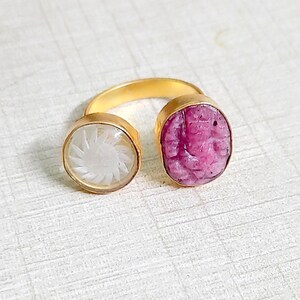 May include: Gold ring with two round stones. The left stone is white and has a carved flower design. The right stone is pink and has a carved design.