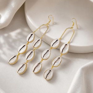 May include: A pair of gold-toned dangle earrings featuring a U-shaped metal wire frame. Each earring is adorned with three white cowrie shells suspended in a vertical line, attached by small gold-colored metal jump rings and a hook-style closure.