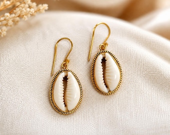 May include: A pair of drop earrings featuring white cowrie shells set in gold-toned metal frames with a twisted rope texture. The earrings hang from simple gold-toned wire hooks, showcasing the natural oval shape and ribbed center of the shells.