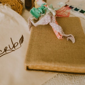 May include: A beige burlap-covered journal with a colorful ribbon tied around it. A cream-colored bag with the word "scrib" and a feather logo is to the left. A watercolor paint set and pencil are in the background.