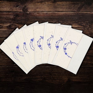 May include: A set of cream-coloured stationery cards featuring a blue floral wreath design. The cards are fanned out, with a white envelope visible. The background is a dark brown wooden surface.