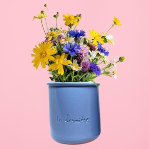 May include: A blue ceramic vase with the text "La fermière" holds a vibrant bouquet of wildflowers. The arrangement includes yellow, blue, purple, and white blossoms. The vase is cylindrical and sits against a pink background.