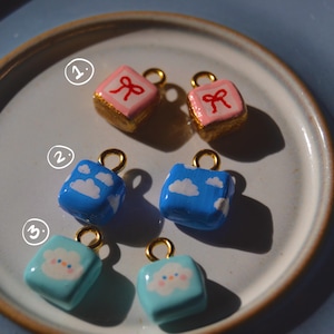 May include: A collection of handmade cube-shaped charms with gold-colored loops. The charms feature various designs: pink with red bows, blue with white clouds, and light blue with a smiling cloud face. The charms are displayed on a white plate with a brown rim.