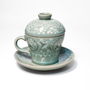 Könnte beinhalten: Ein hellgrünes Keramik-Teetassen- und Untertassen-Set mit Deckel. Tasse und Untertasse sind mit weißen Blumen- und Vogelmotiven verziert.