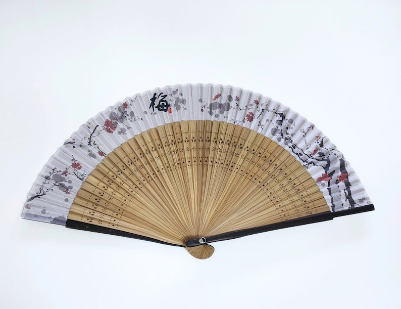 May include: A folding hand fan with a bamboo frame and a white paper panel. The panel features a floral design with red and gray accents, and a black Japanese character. The fan is open, displaying its full shape.