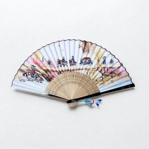 May include: A decorative folding fan with a painted landscape scene. The fan features a light blue background with a detailed illustration of figures on horseback, a carriage, and trees. The fan has a wooden handle and a blue tassel.