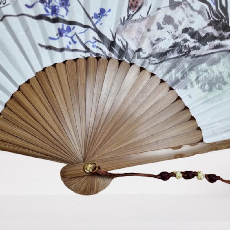 May include: A hand fan with a bamboo frame and a painted paper panel. The paper features a watercolor landscape with blue flowers and a bird. A brown cord with decorative knots hangs from the fan's base.