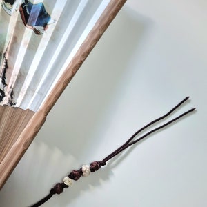 May include: A white paper folding fan with a bamboo frame. The fan features a painted design with figures in traditional clothing. A dark brown tassel with decorative beads hangs from the fan.