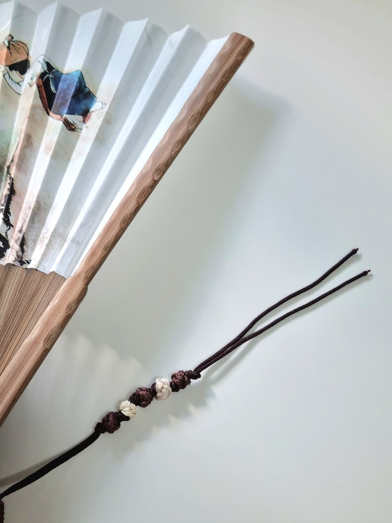 May include: A white paper folding fan with a bamboo frame. The fan features a painted design with figures in traditional clothing. A dark brown tassel with decorative beads hangs from the fan.