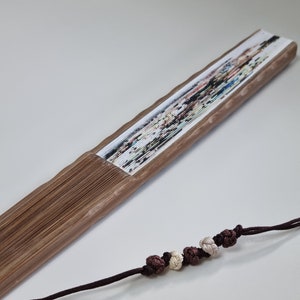 May include: A wooden folding fan with a colorful floral design. The fan is brown and has a decorative cord with knotted beads in shades of brown and cream. The fan is partially open, displaying the printed artwork.