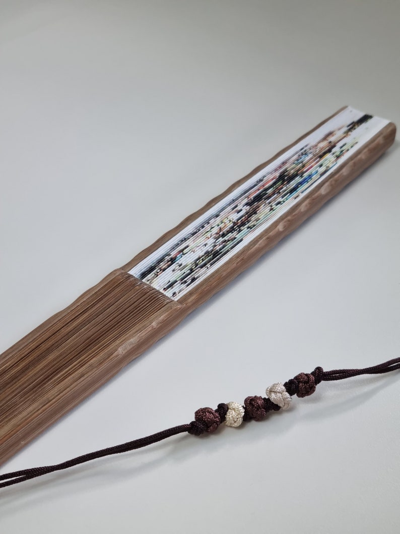 May include: A wooden folding fan with a colorful floral design. The fan is brown and has a decorative cord with knotted beads in shades of brown and cream. The fan is partially open, displaying the printed artwork.