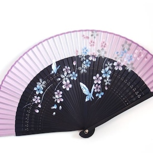May include: A decorative folding fan with a black wooden frame and a light purple and white fabric. The fan features a floral design with butterflies in shades of blue and pink. The fan is open, displaying its full design.