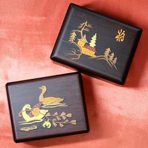 Op de afbeelding: Twee houten dozen met ingelegde ontwerpen. De bovenste doos heeft een pagode en bomen, met het Chinese karakter "猫" (kat) op de deksel. De onderste doos heeft een paar ganzen die in een vijver zwemmen.