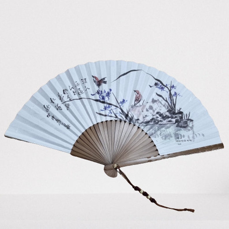 May include: A light blue, hand-painted fan with a landscape scene featuring birds, trees, and mountains. The fan has a wooden handle and a decorative tassel. Calligraphy is visible on the fan's surface.