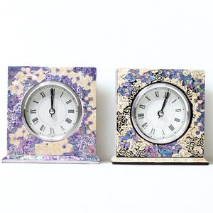 May include: Two square decorative clocks with floral designs in purple, cream, and black. Each clock has a silver-coloured face with black Roman numerals and hands. The clocks are on a white surface.