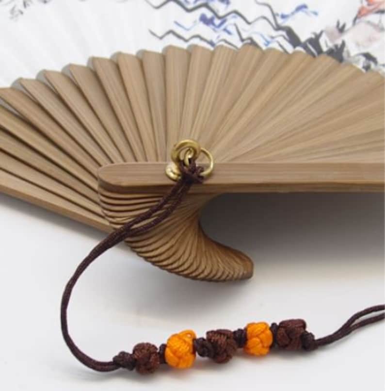 May include: A close-up of a bamboo hand fan, partially open, with a decorative cord. The fan features light brown wooden slats and a detailed, carved handle. The cord is dark brown with orange and brown decorative knots.