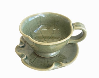May include: A green ceramic teacup and saucer set with a leaf design. The cup has a rounded shape and a handle, while the saucer has a scalloped edge.