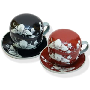 May include: Two ceramic teacups with lids and matching saucers. The cups are decorated with white floral designs on a black and red background.