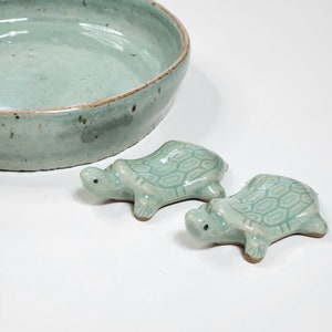 May include: Two small, green ceramic turtle figurines. The turtles are facing the right side of the image. The turtles are sitting on a white surface.