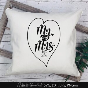 May include: White square pillow with a black heart-shaped design. The design includes the words "Mr. and Mrs. est. 2021". The pillow is on a wooden surface with a decorative frame and greenery.