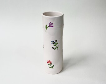 Könnte beinhalten: Weiße Keramikvase mit zylindrischer Form und floralem Design. Die Vase ist mit kleinen, handbemalten Blumen in den Farben Rosa, Blau, Gelb und Grün verziert.