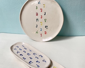 May include: A set of three off-white ceramic serving dishes. The largest is round, the other two are oval. Each dish features colorful, abstract symbols. The background is a light blue wall.