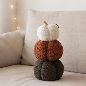 May include: A stack of three plush pumpkins made of textured sherpa fabric. The top pumpkin is cream, the middle is rust orange, and the bottom is dark olive green. Each pumpkin has a rounded, segmented shape with a small tan stem on the top one.