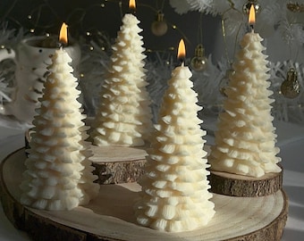 May include: Four ivory-colored, tree-shaped candles with lit flames sit on wooden slices. The candles have a layered design, resembling a Christmas tree. A white Christmas tree with gold ornaments is in the background.